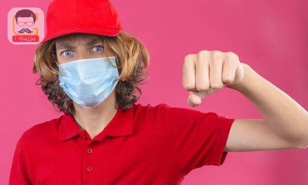 young delivery man in red uniform wearing medical mask showing fist to camera aggressive facial expression over isolated fist background