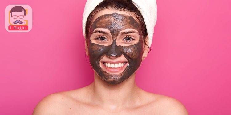 Excited happy female poses with toothy smile and chocolate facial mask, with nacked shoulders, takes care of her beauty and appearance, wears white towel on head, isolated over pink background.